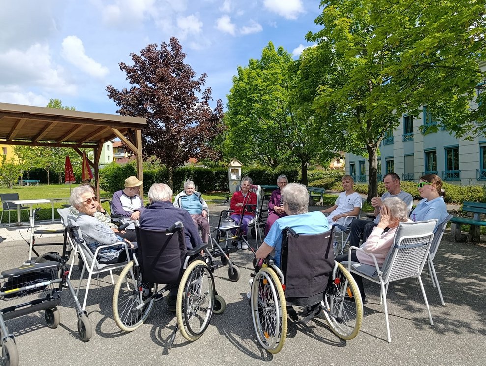 BewohnerInnen, Praktikantin und Mann (FSJ) sitzen im Rollstuhl oder auf Sessel im Garten bei der Morgenrunde.