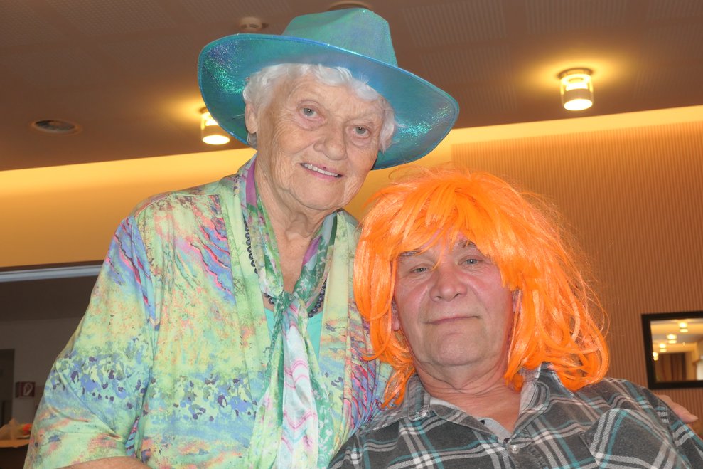 Eine Dame mit Hut und ein Herr mit oranger Perücke.