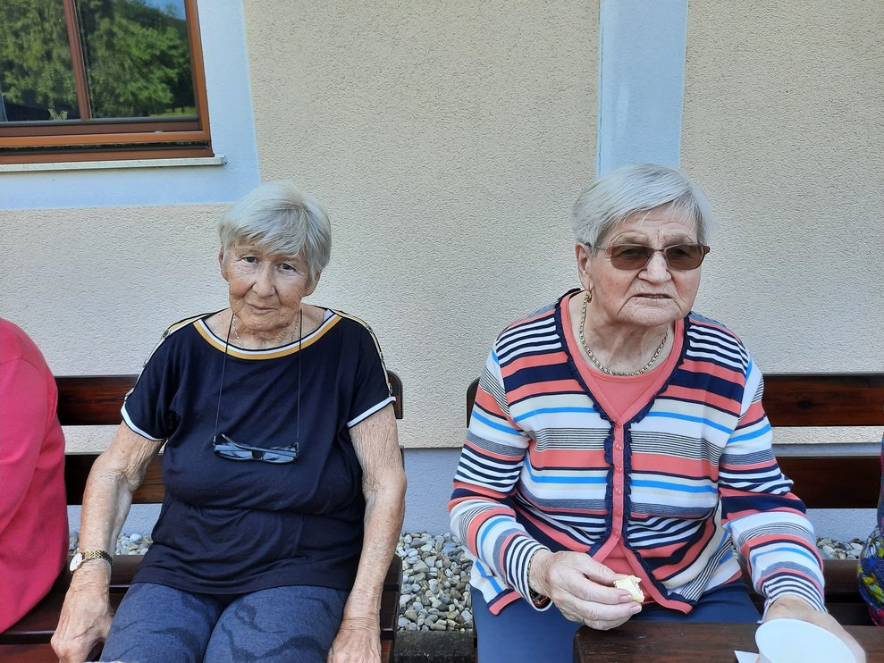 Zwei Damen sitzen auf einer Bank, vor einem Haus.