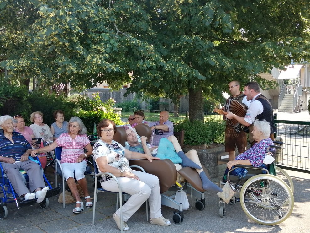 Einige Personen sitzen im Schatten der Bäume, die Musiker spielen auf.
