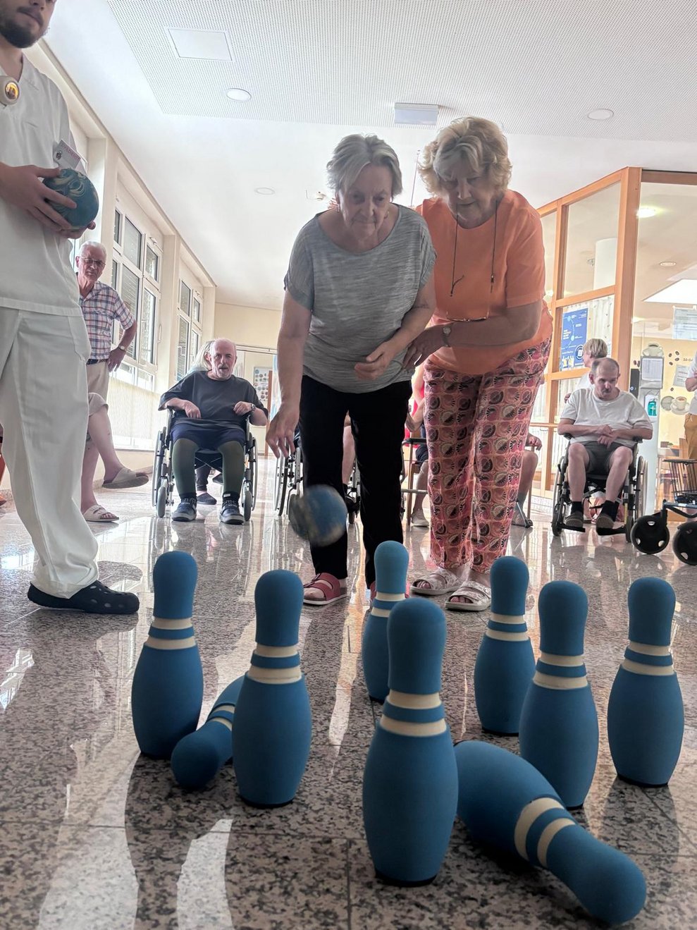 Stehende Bewohnerin beim Kegeln. Kegelball in der Luft. Ehrenamtlich Tätige hält Bewohnerin.