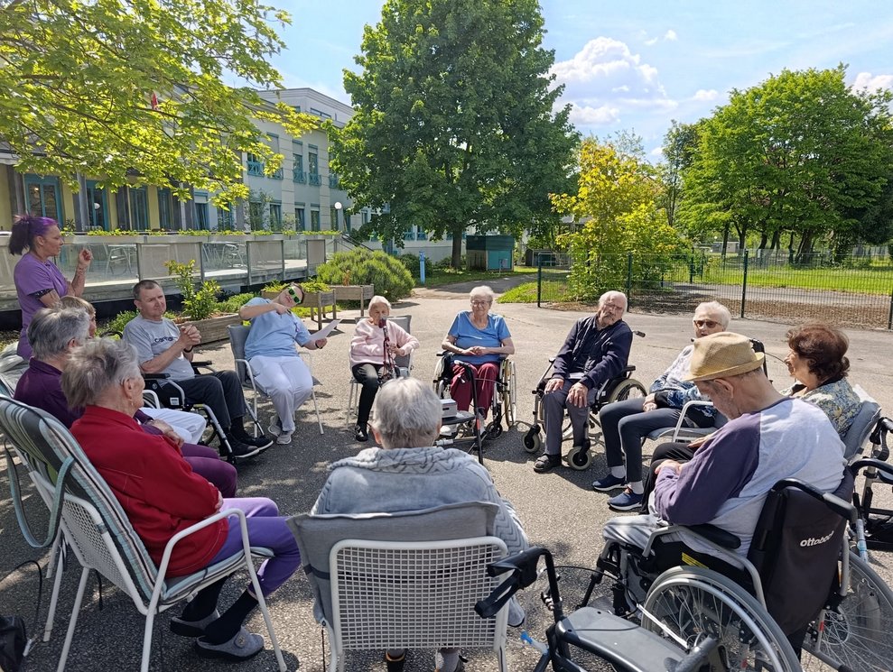 BewohnerInnen, Praktikantin und Mann (FSJ) sitzen im Rollstuhl oder auf Sessel im Garten bei der Morgenrunde. Alltagsbegleiterin steht.