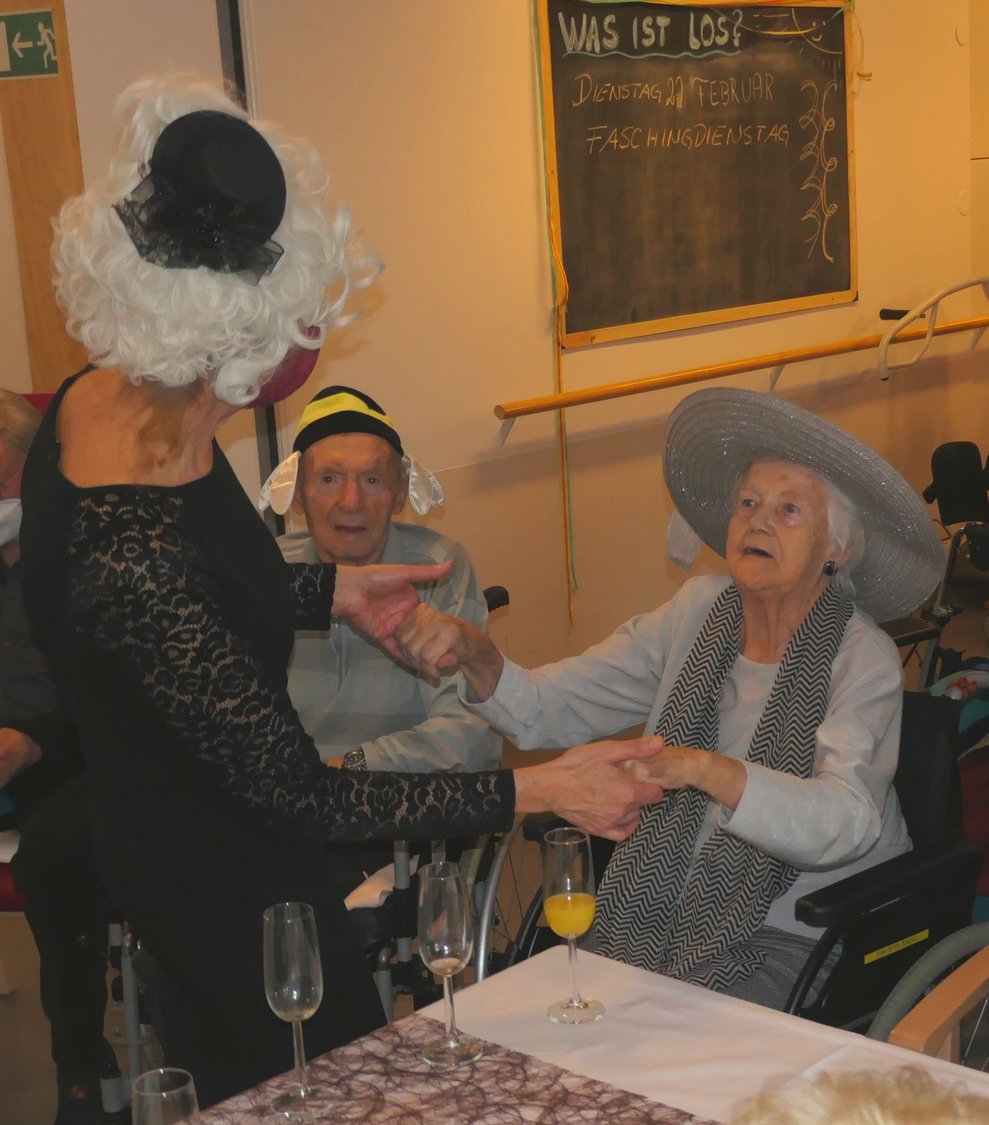 Eine Dame im Ballkleid fordert eine Dame mit großem Hut zum Tanzen auf.