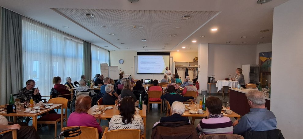 Viele sitzende Personen. Auf Leinwand wird Foto und Text gezeigt.