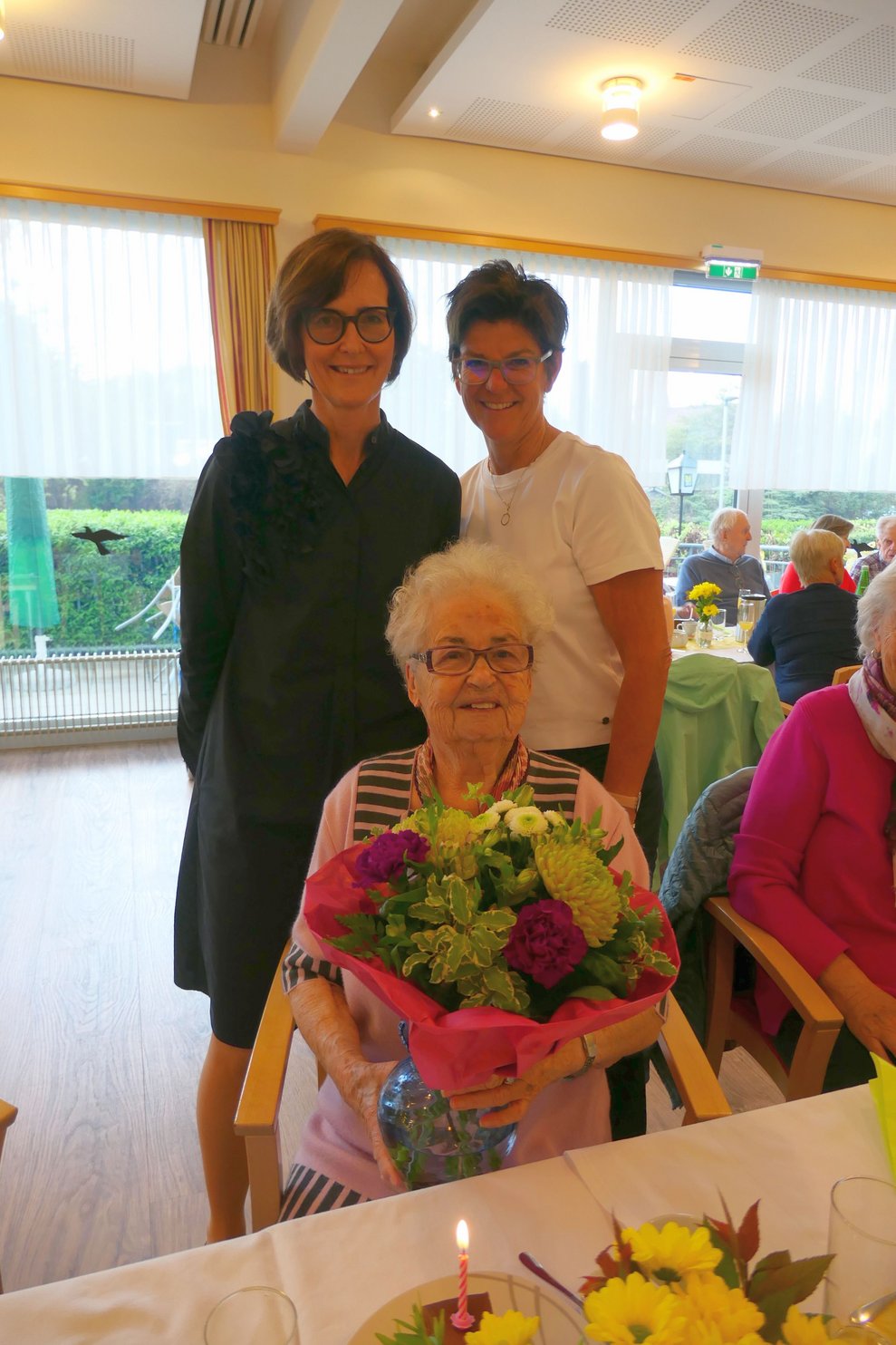 Zwei Damen gratulieren Bewohnerin mit Blumenstrauß.