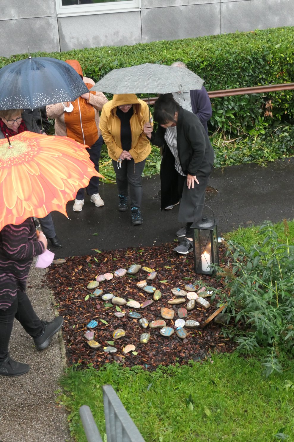 Einige Personen mit Regenschirmen vor einem Quadrat am Gartenboden, in welchem aus Steinen ein Herz und Blumen gelegt sind.
