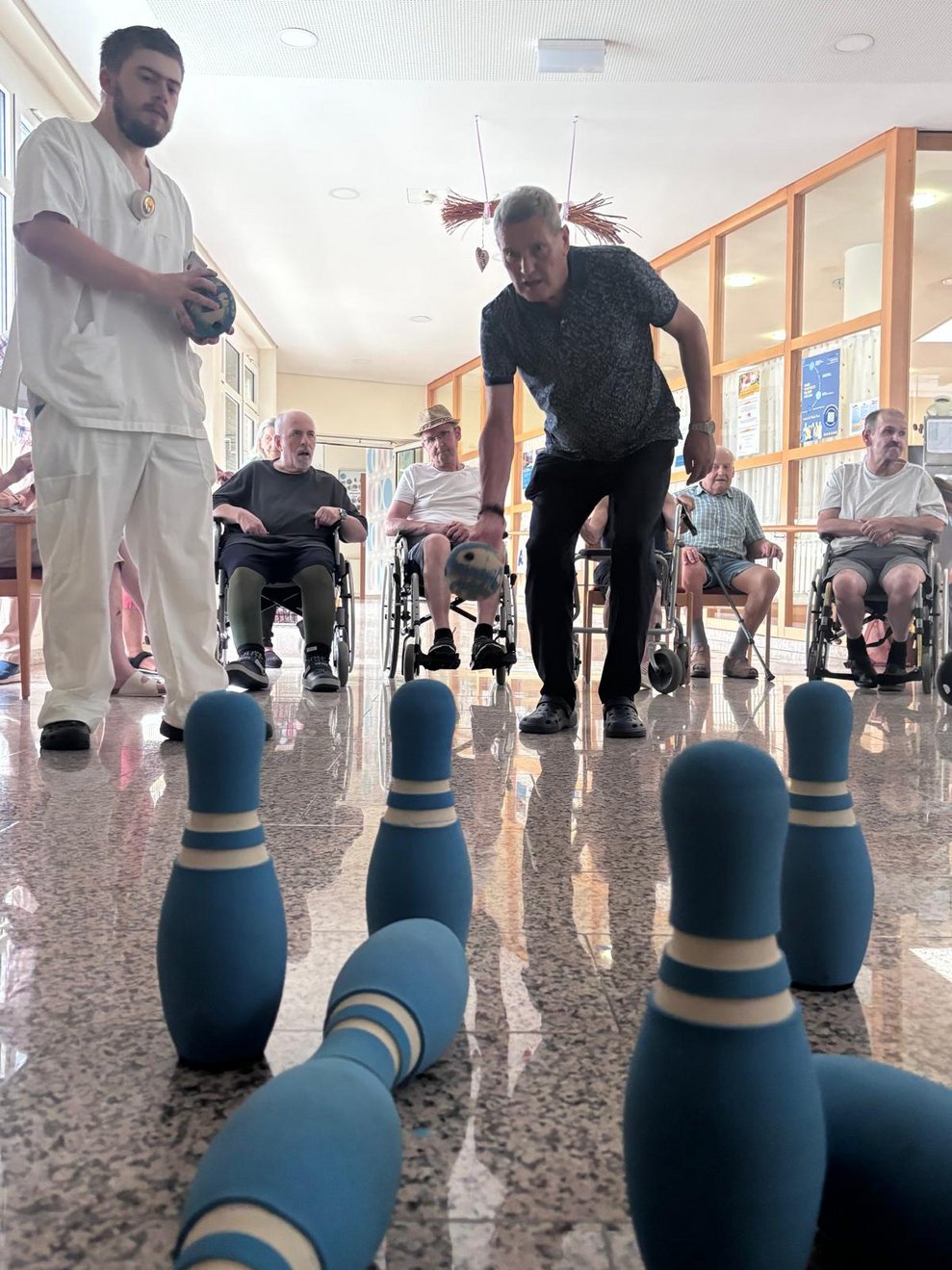 Stehende Bewohnerin beim Kegeln. Bewohner zielt mit Kegelball auf die Kegel. Paar Kegel sind umgefallen. Mitarbeiter hält Kegelball in den Händen.