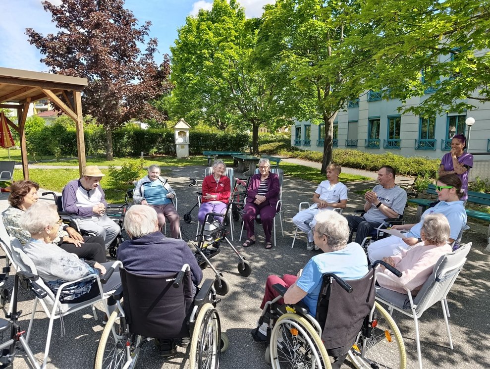 BewohnerInnen sitzen im Rollstuhl oder auf Sessel im Garten bei der Morgenrunde.
