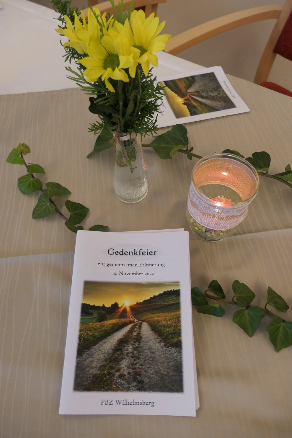 Ein Tischtuch, gelbe Blumen, Teelicht im Glas, Efeuranken und ein Programmheft.