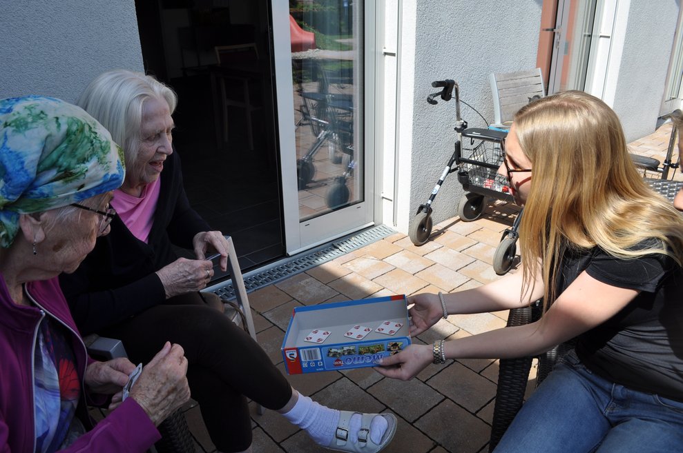 ein junges Mädchen lässt zwei ältere Damen eine Spielkarte aus einer Schachtel ziehen