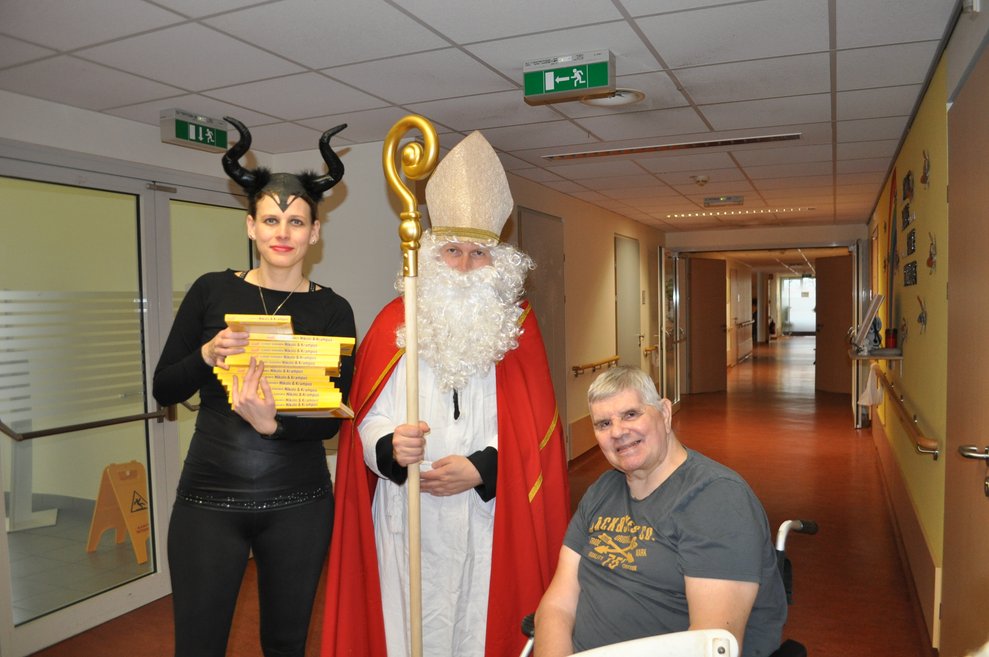 eine Frau als Krampus verkleidet und ein Mann als Nikolaus verkleidet zu Besuch bei einem Herrn im Pflegeheim