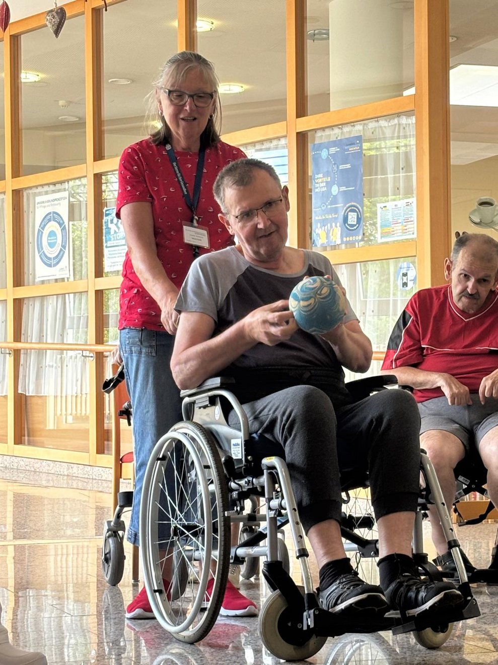 Bewohner hält Kegelball in den Händen und ehrenamtlich Tätige hält Rollstuhl mit Bewohner.