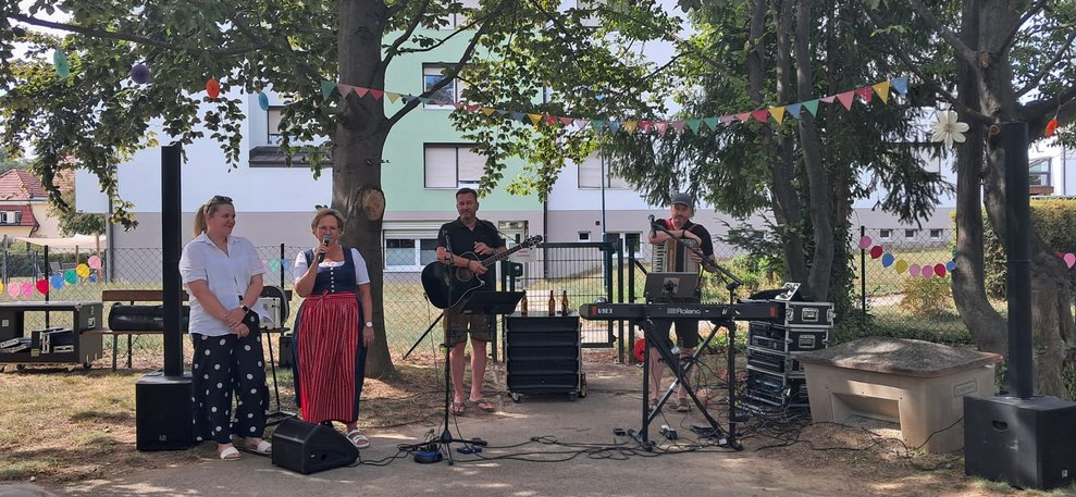 Frau redet ins Mikrofon. Frau steht daneben. Liveband zwei Männer mit Instrumenten.