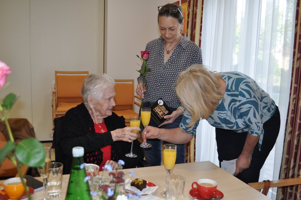eine Dame gratuliert einer älteren Dame zum Geburtstag und eine weitere Dame hält eine Rose in der Hand