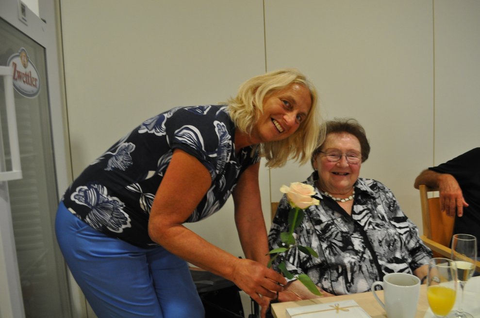 eine ältere Dame bekommt von einer Frau Blumen und Pralinen zum Geburtstag überreicht