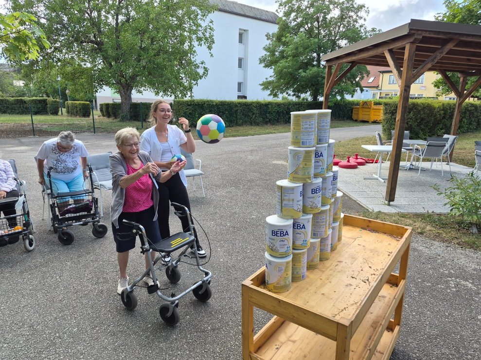 Bewohnerin beim Dosenschießen. Ball in der Luft. Praktikantin steht daneben und hält Ball in der Hand.