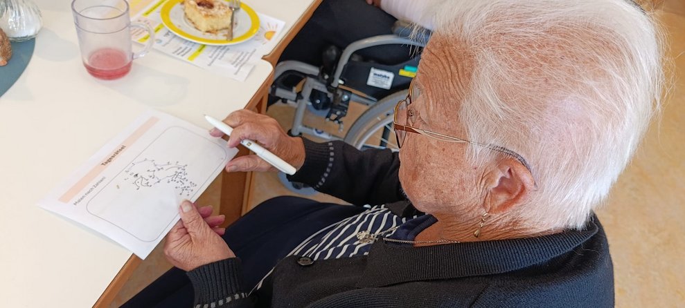 Bewohnerin hält Stift in der Hand. Morgenpost liegt auf Tisch.