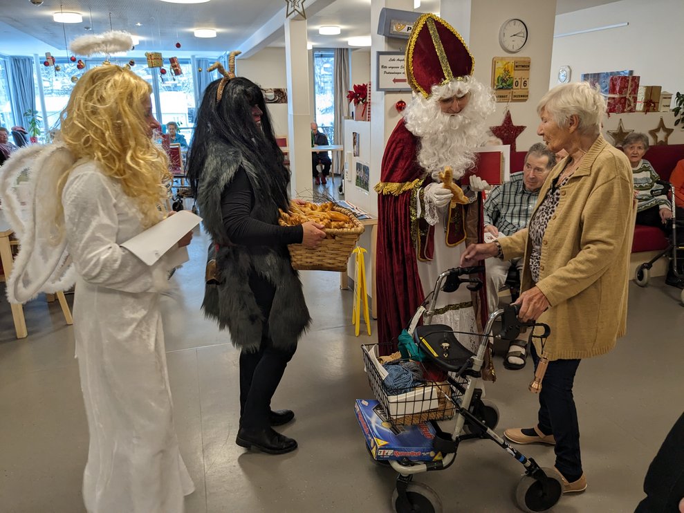 Der Nikolo übergibt ein Geschenk, Krampus trägt den Korb mit den Backwaren.