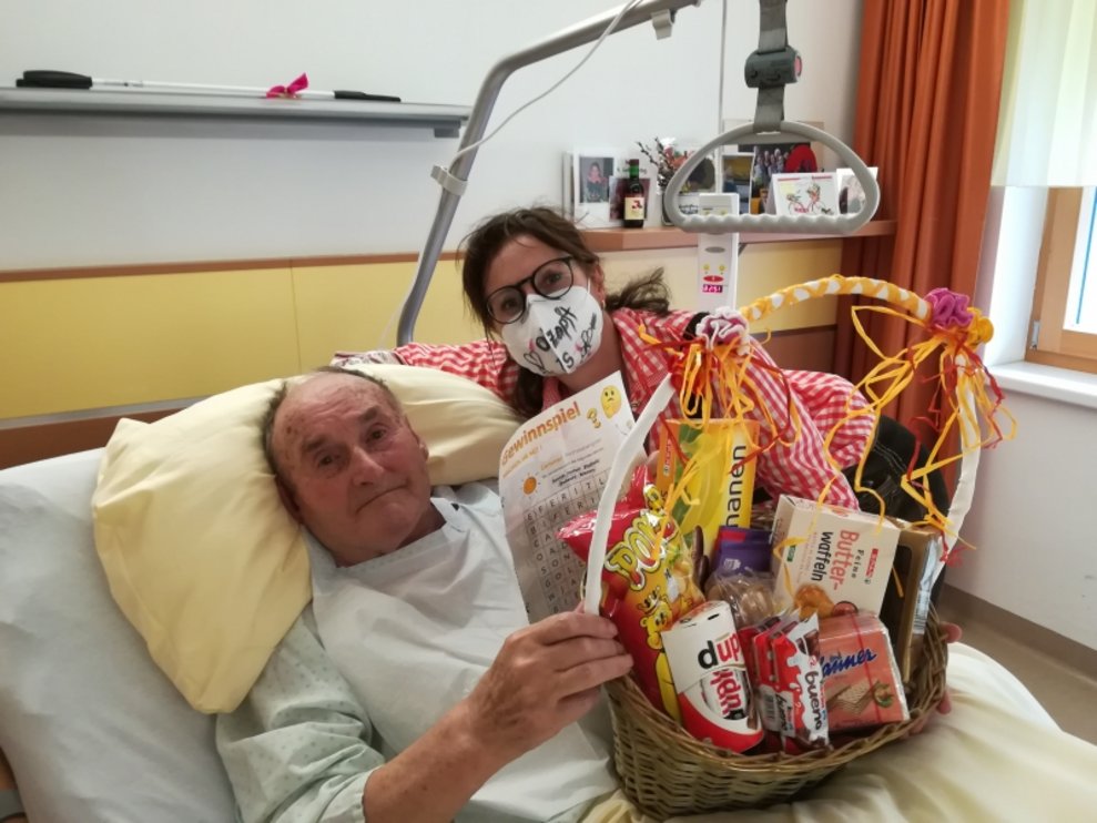 Ein Bewohner liegt mit einem Geschenkkorb im Bett und eine Betreuerin steht neben ihm.