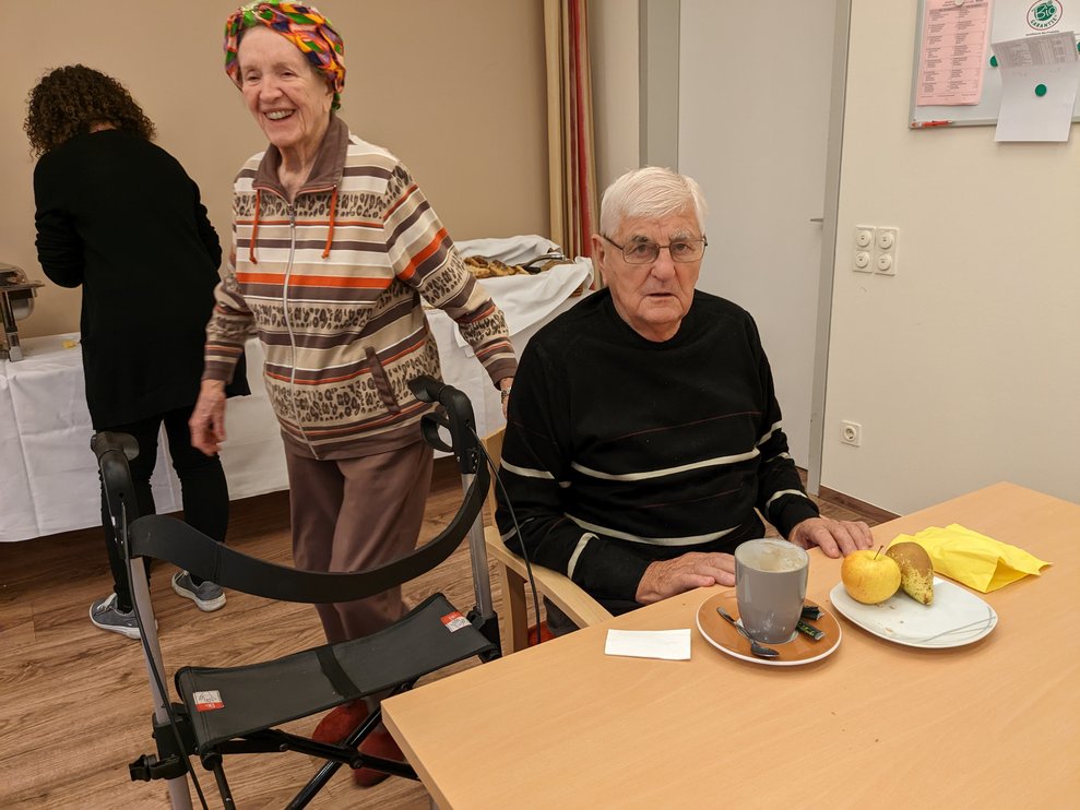Eine Dame mit Rollator und ein Herr bei Tisch.