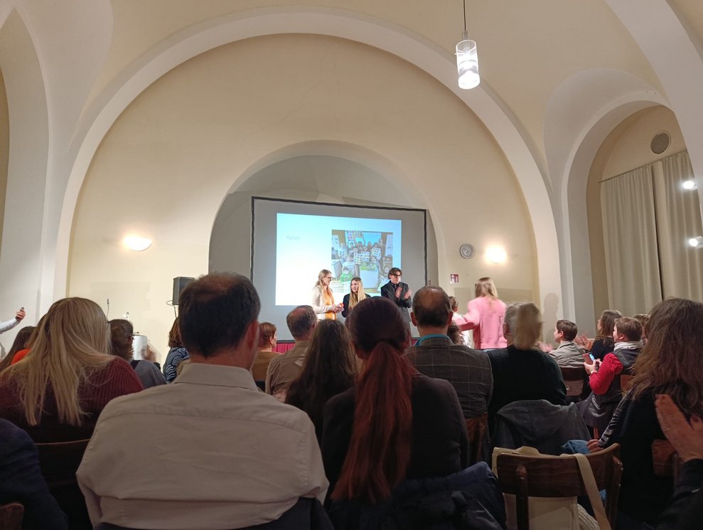 Personen sitzen bei der Präsenation. Schülerinnen und Lehrerin stehen auf Bühne.