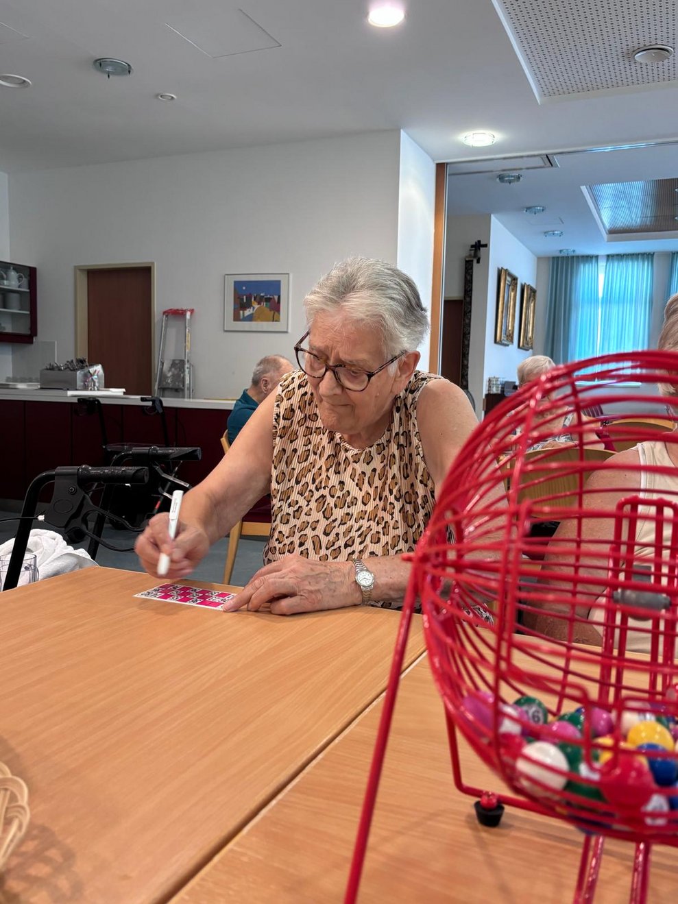 Bewohnerin beim Bingo spielen und hält Stift in der Hand. Zahltafel liegt auf Tisch. Bingortrommel steht auf Tisch.