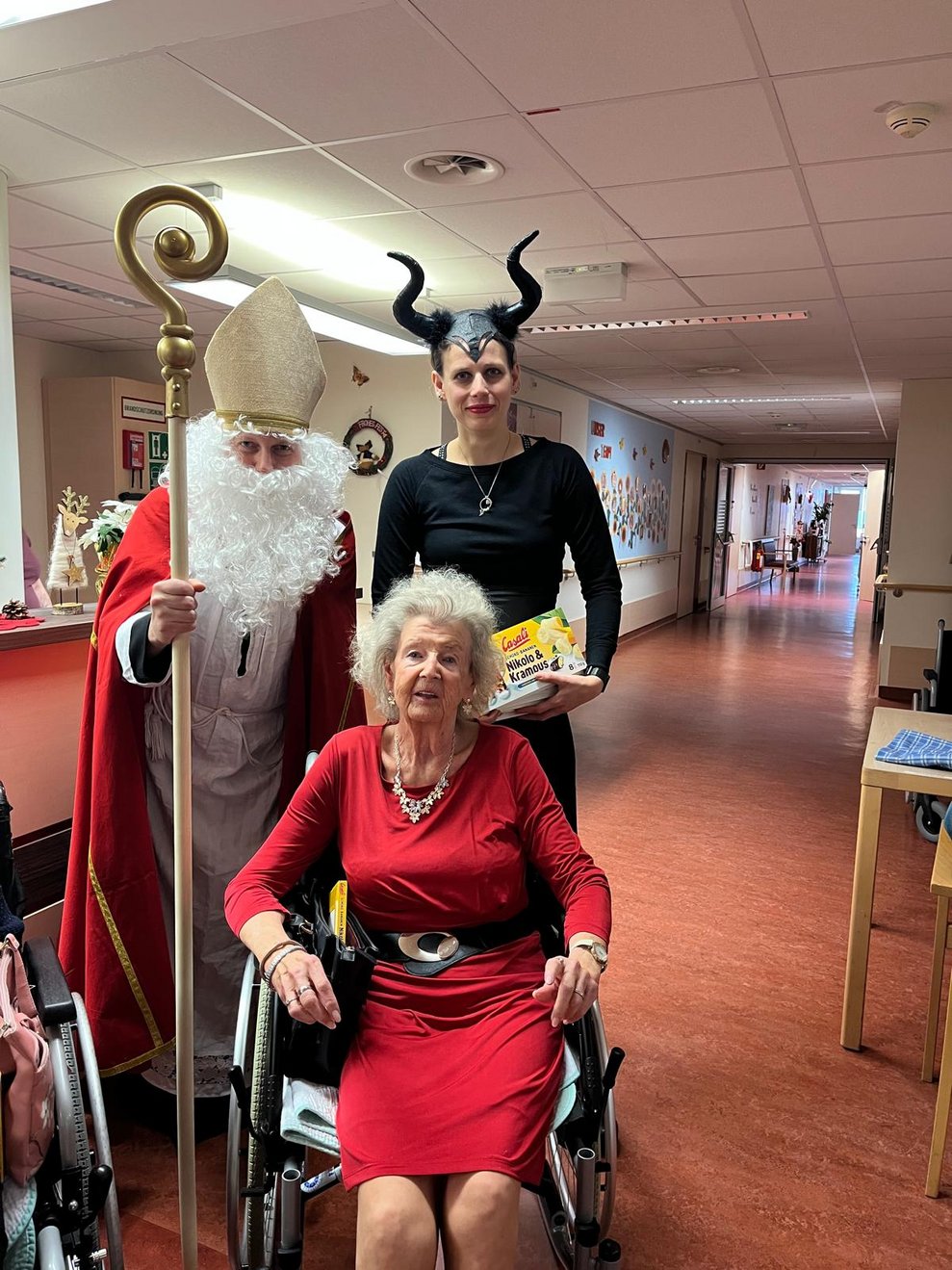 eine Frau als Krampus verkleidet und ein Mann als Nikolaus verkleidet zu Besuch bei einer älteren Dame im Pflegeheim
