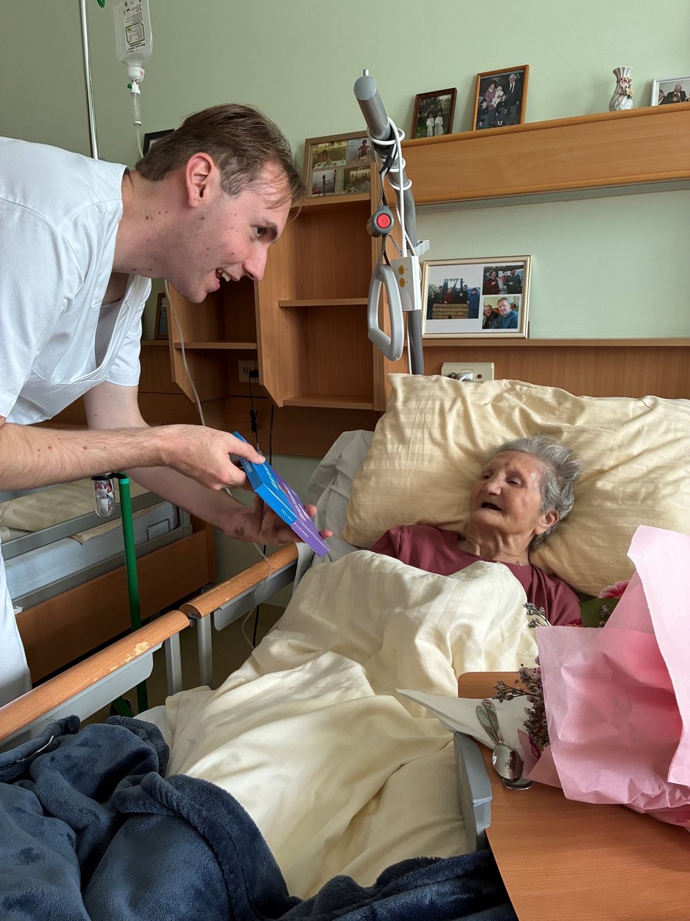 Alltagsbegleiter hält Milka Pralinen Packung in der Hand und gratuliert liegende Bewohnerin im Bett zum Geburtstag.