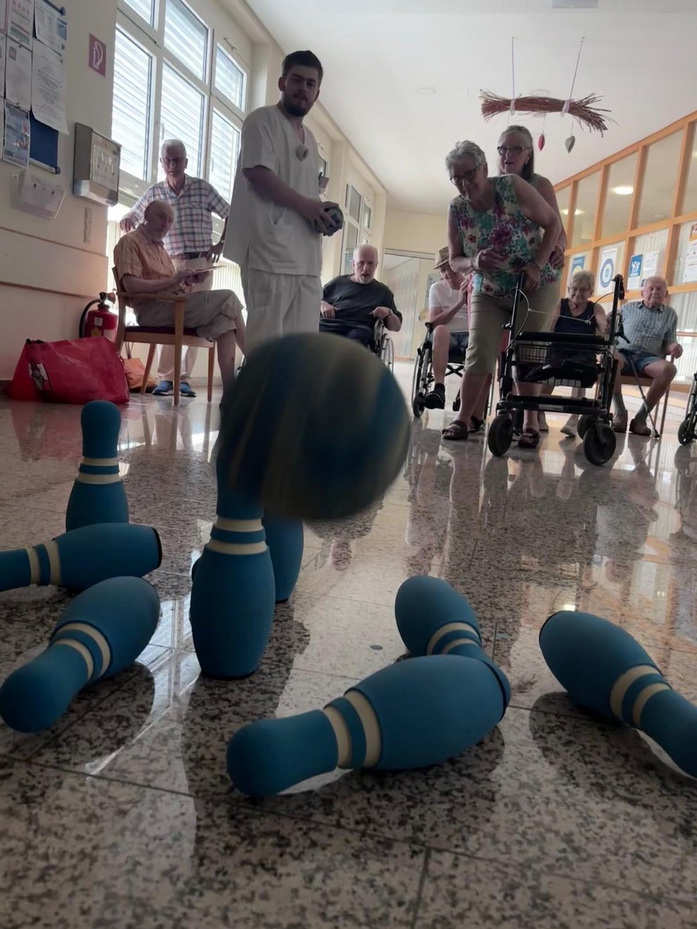 Stehende Bewohnerin beim Kegeln. Ehrenamtlich Tätige hält Bewohnerin. Mitarbeiter hält Kegelball in den Händen. Kegelball in der Luft. Paar Kegel sind umgefallen.