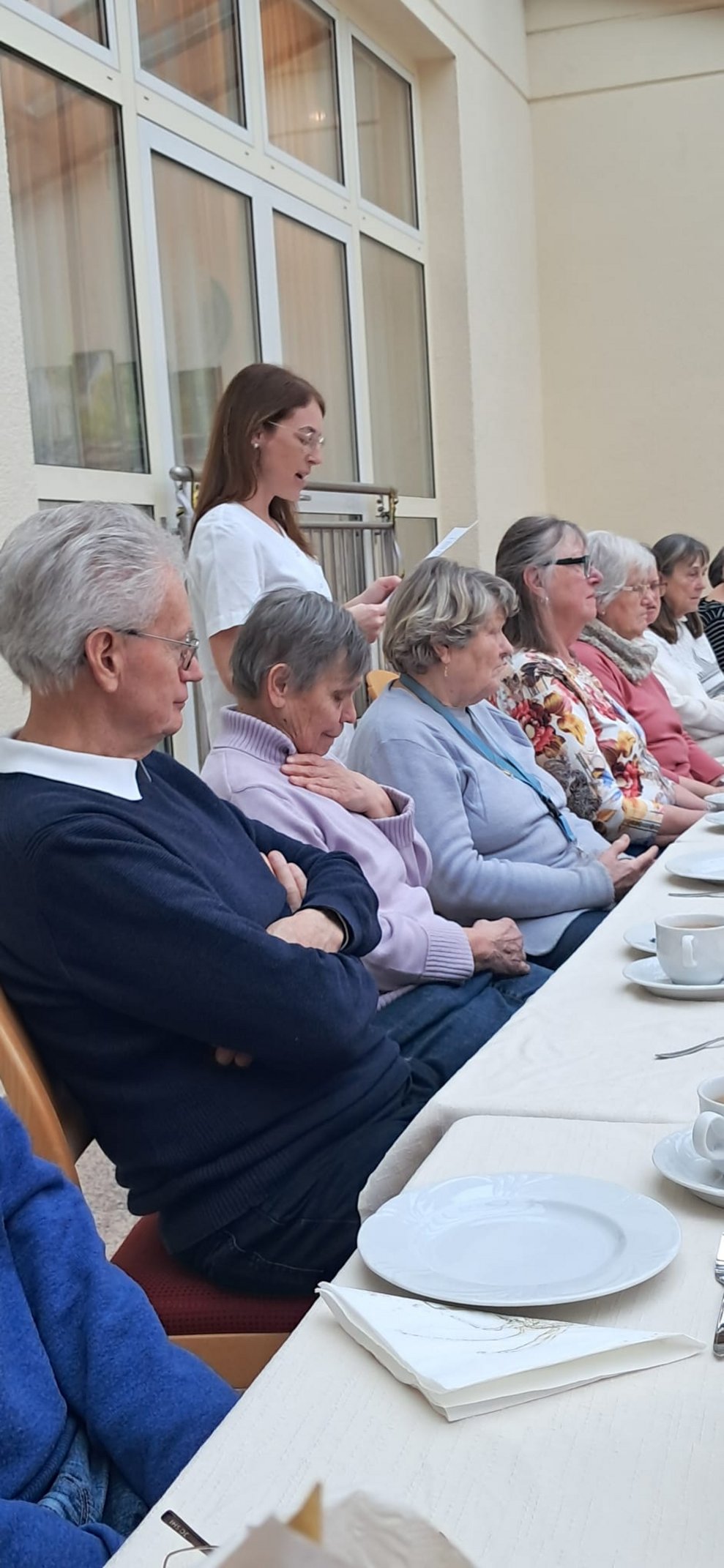 Alltagsbegleiterin ließt vom Zettel. Viele Personen sitzen.