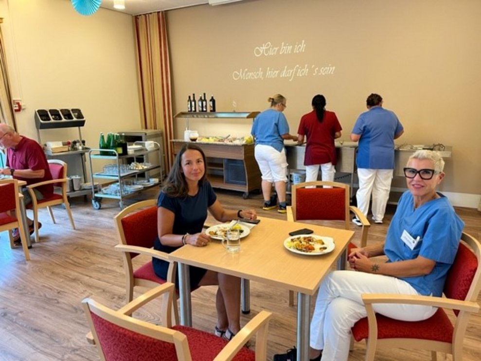 2 Damen bei Tisch, im Hintergrund sind 3 Damen beim Buffet.