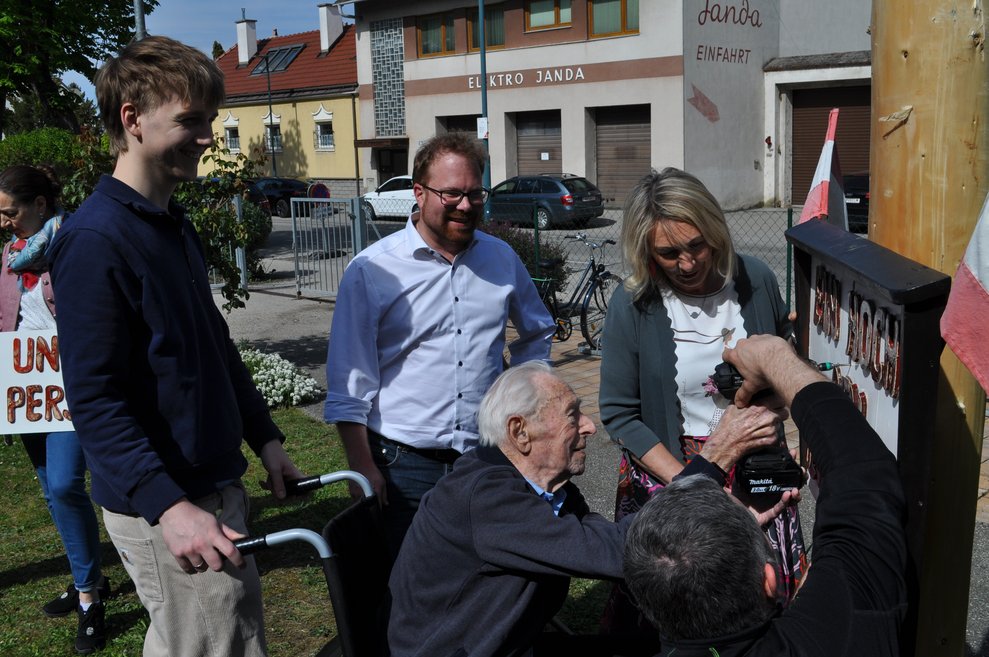 ein älterer Herr bohrt mit Hilfe ein Schild an den Maibaum,