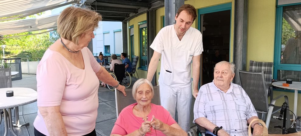 Bewohnerin häkelt. Frau und Alltagsbegleiter stehen und Bewohner sitzt.