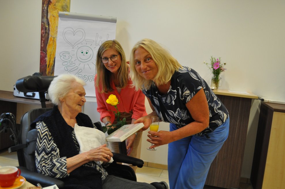 Zwei Damen gratulieren einer älteren Dame im Rollstuhl zum Geburtstag