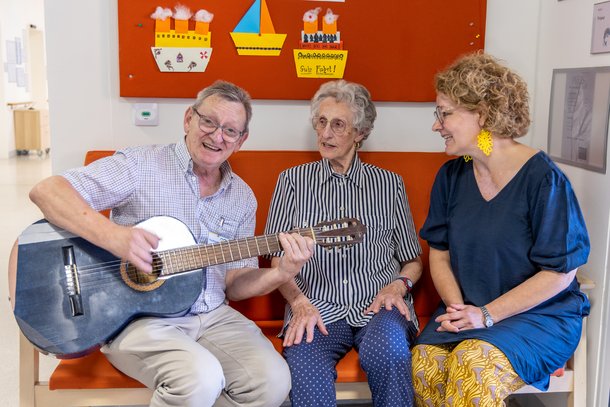 Ehrenamtlicher spielt vor Landesrätin Teschl-Hofmeister und einer Bewohnerin mit der Gitarre