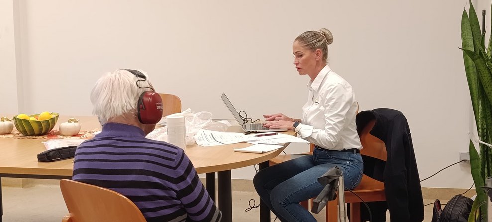 Sitzender Bewohner hat Kopfhörer beim Hörtest auf. Frau sitz auf Sessel und bedient Computer. 