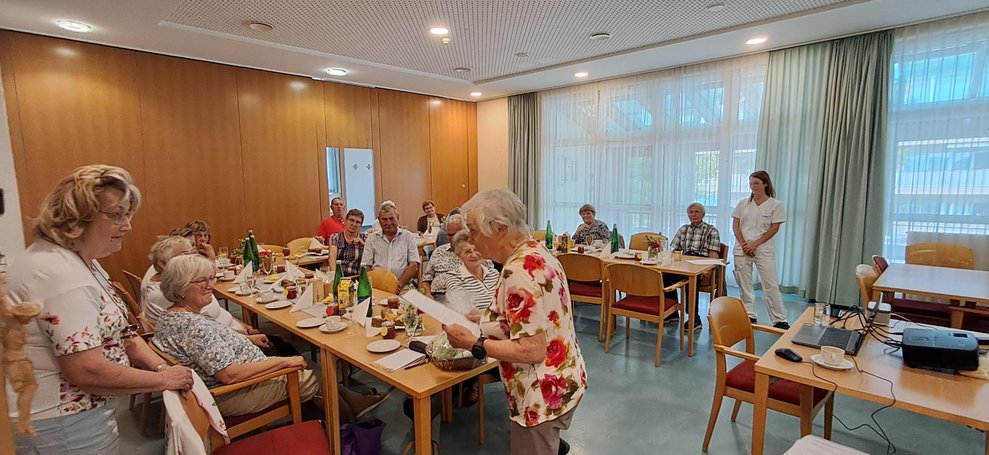 Ehrenamtlich Tätige steht, hält Zettel in der Hand und liest. Viele sitzende Personen. Alltagsbegleiterin steht. Eine ehrenamtlich Tätige und Alltagsbegleiterin stehen.