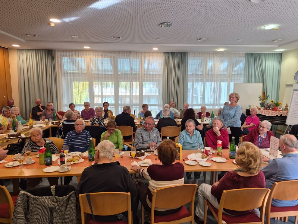 Frau steht und redet. Sitzende Personen beim Tisch.