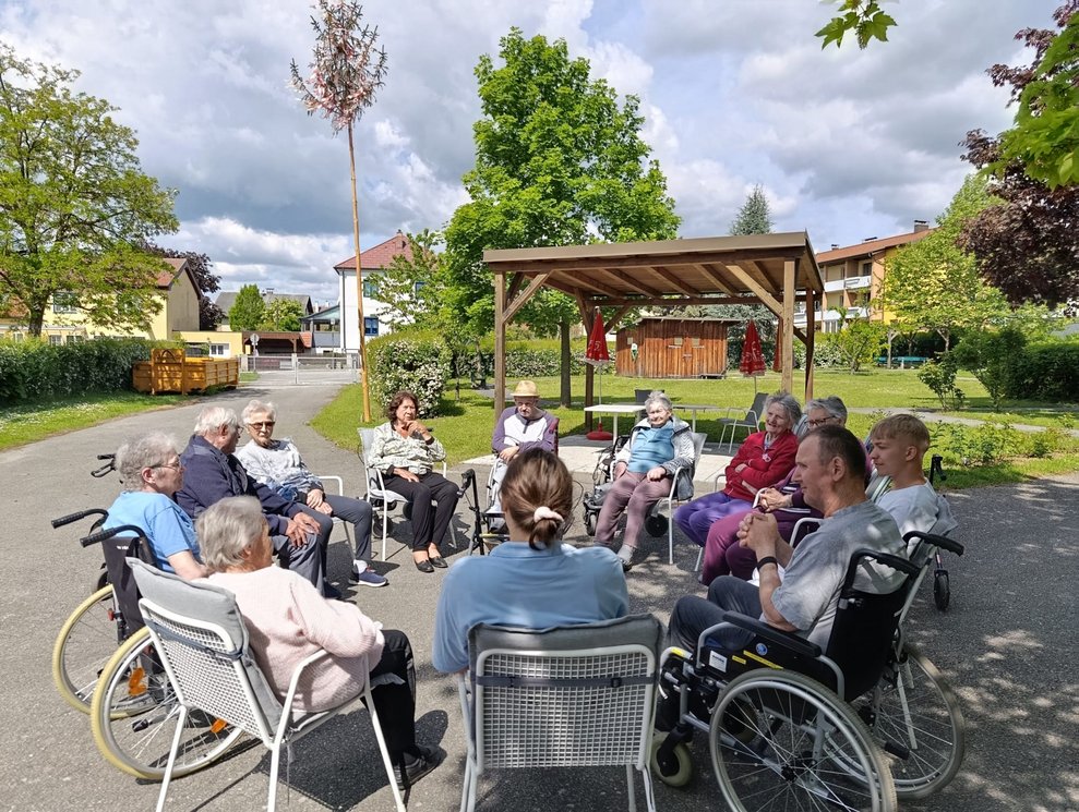 BewohnerInnen, Praktikantin und Mann (FSJ) sitzen im Rollstuhl oder auf Sessel im Garten bei der Morgenrunde.