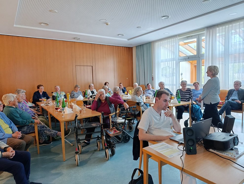 Viele BewohnerInnen bei der BewohnerInnenbesprechung. Mann sitzt beim Computer. Frau redet.