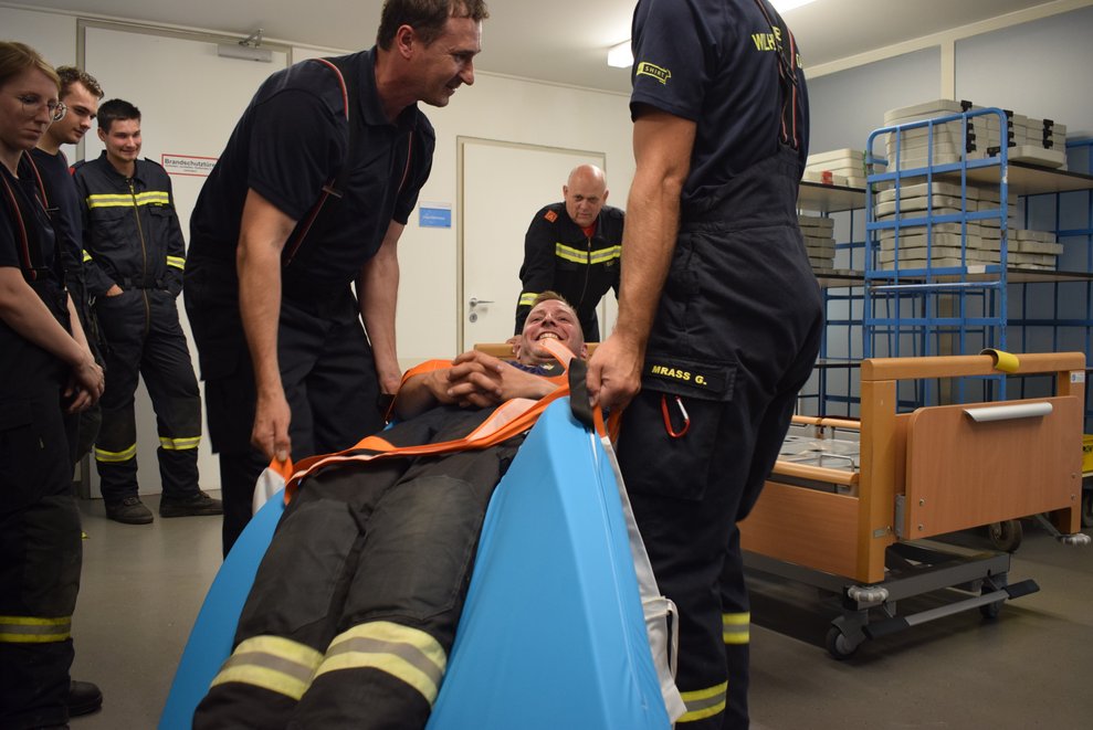 Der Feuerwehrmann wird von zwei Kollegen mit der Evakuierungsmatte aus dem Bett gehoben.
