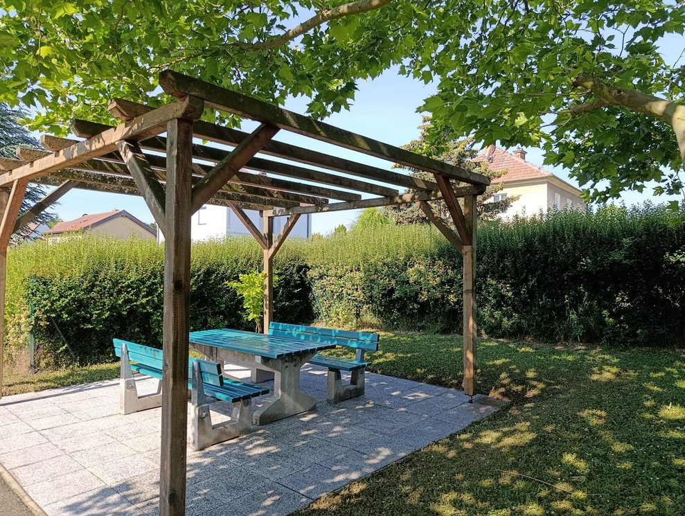 Platz mit Gartenbänke und Tisch. Darüber ein offenes Holzdach mit Gestänge.