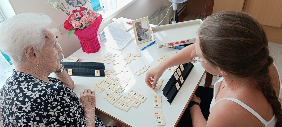 Bewohnerin und Praktikantin spielen Rummikub. Spielsteine liegen auf Tisch.