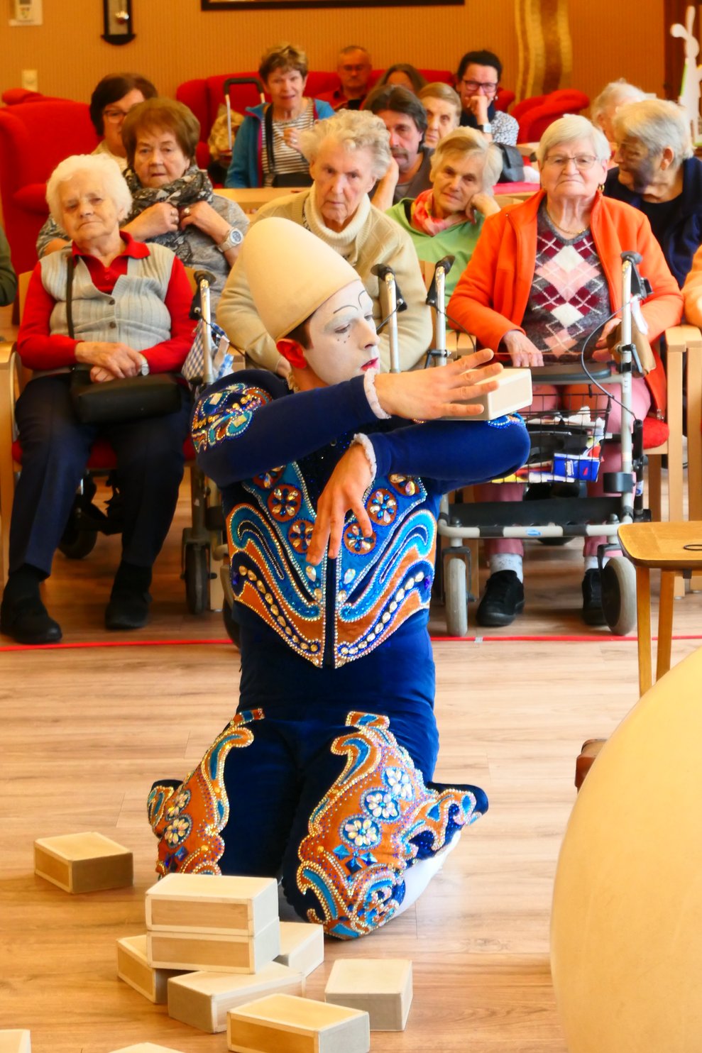 Der Clown kniet auf dem Boden und legt sich einen Holzblock auf die Schulter.