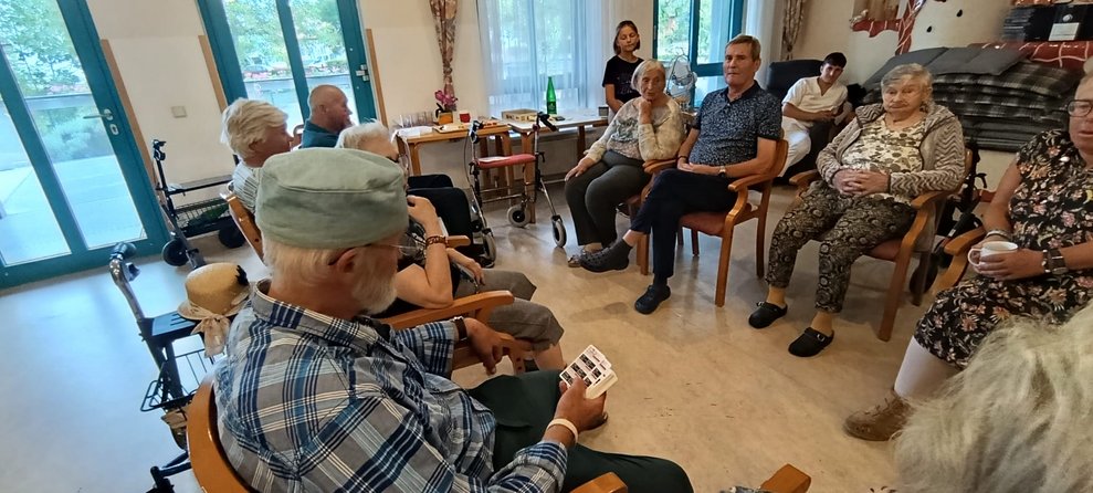 Mann (ehrenamtlich Tätiger) hält Quizkarten in der Hand. Sitzende BewohnerInnen.