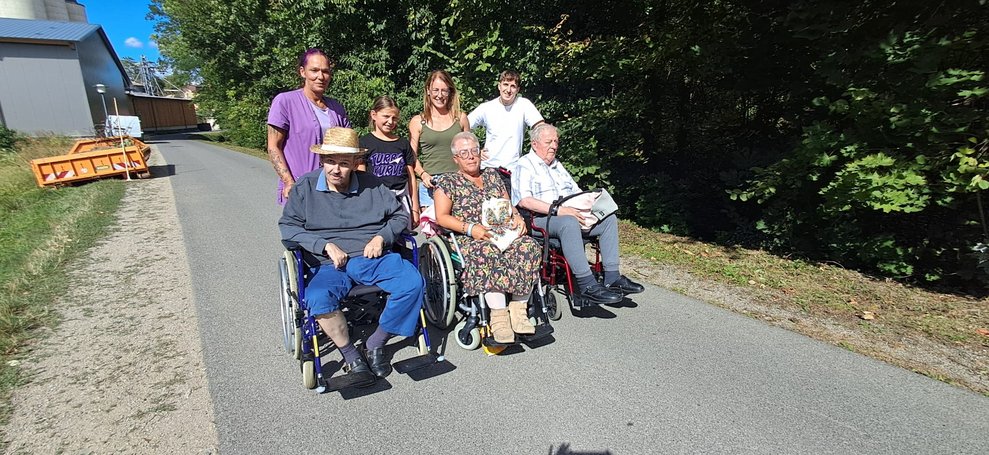 BewohnerInnen im Rollstuhl. Personen halten Rollstuhl bei den Griffen.