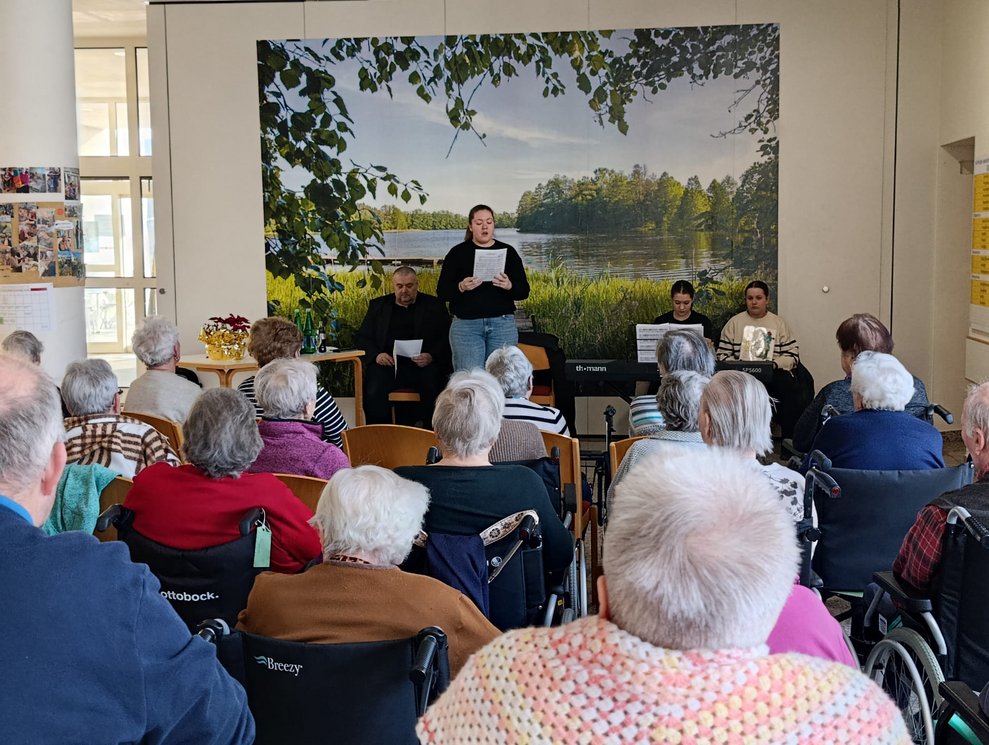 Viele BewohnerInnen im Rollstuhl und Personen sitzen auf Sessel. Mädchen singt.
