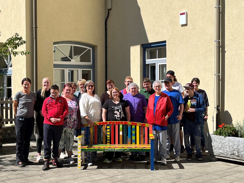 Viele Personen stehen hinter einer bunten Gartenbank.