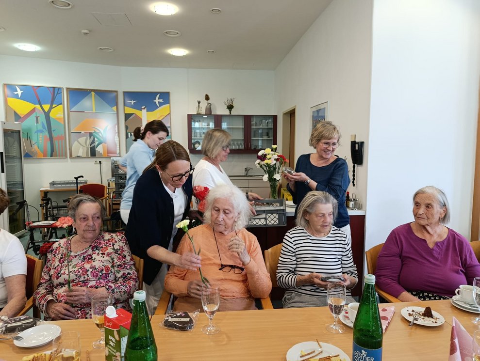 Pflege- und Betreuungsmanagerin gibt Bewohnerin eine Nelke. Bewohnerin mit Nelke in der Hand. Bewohnerinnen schauen zu.